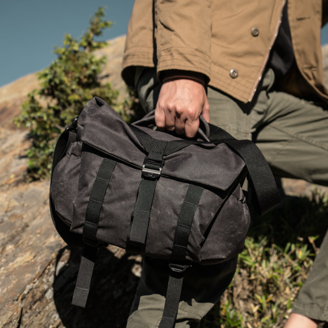 Hand holding the Wotancraft Pilot 7L charcoal bag by its top handle outdoors